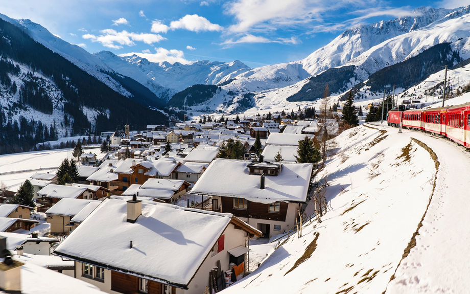 Glacier Express fährt durch eine verschneite Landschaft oberhalb eines sonnigen Dorfes